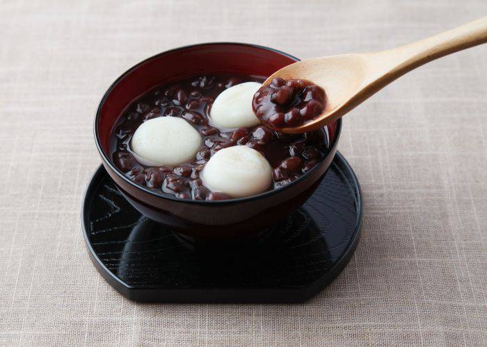 A bowl of Japanese red bean soup, with shiny red beans, chewy mochi, and a wooden spoon.